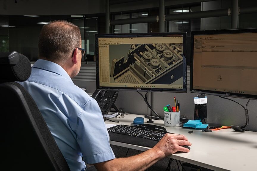 A man focused at a desk, working on two computer screens, surrounded by a tidy workspace.