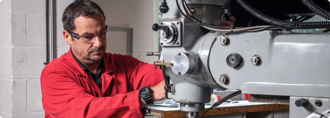 A man in a red shirt operates a machine, focused on his task in a workshop environment.